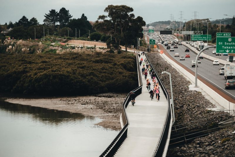 Photo: Long shot of a wooden boardwalk that runs between a motorway and a mangrove forest. The boardwalk is full of people on bikes. 