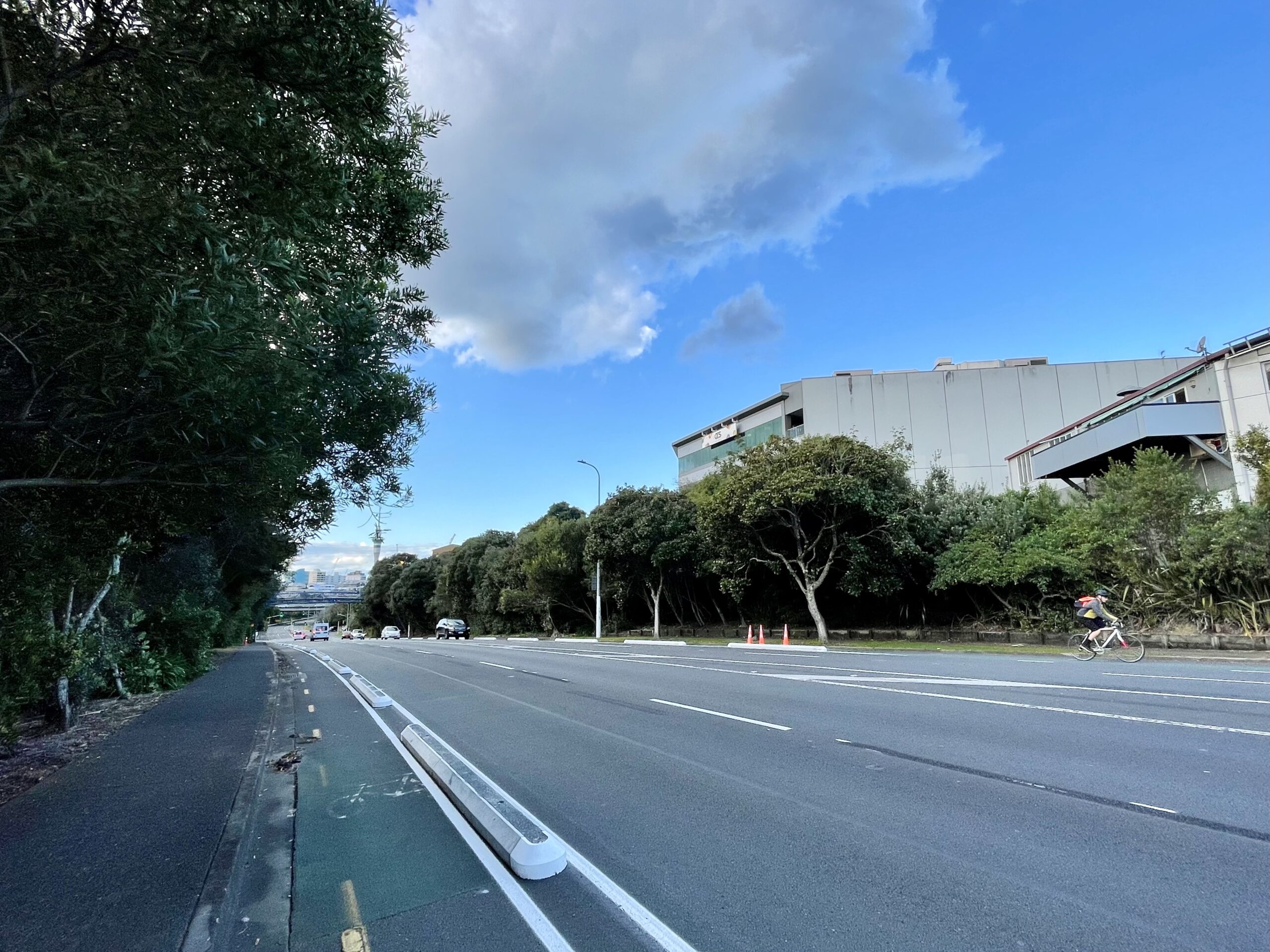 A wide road including a painted cycleway bordered by concrete protectors. Across the road a person is riding a bike.