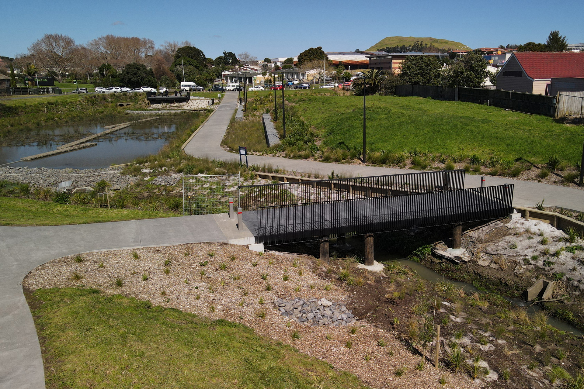 A picturesque reserve, including a small lake, pathways and a bridge. It's surrounded by housing and a maunga is visible in the background.