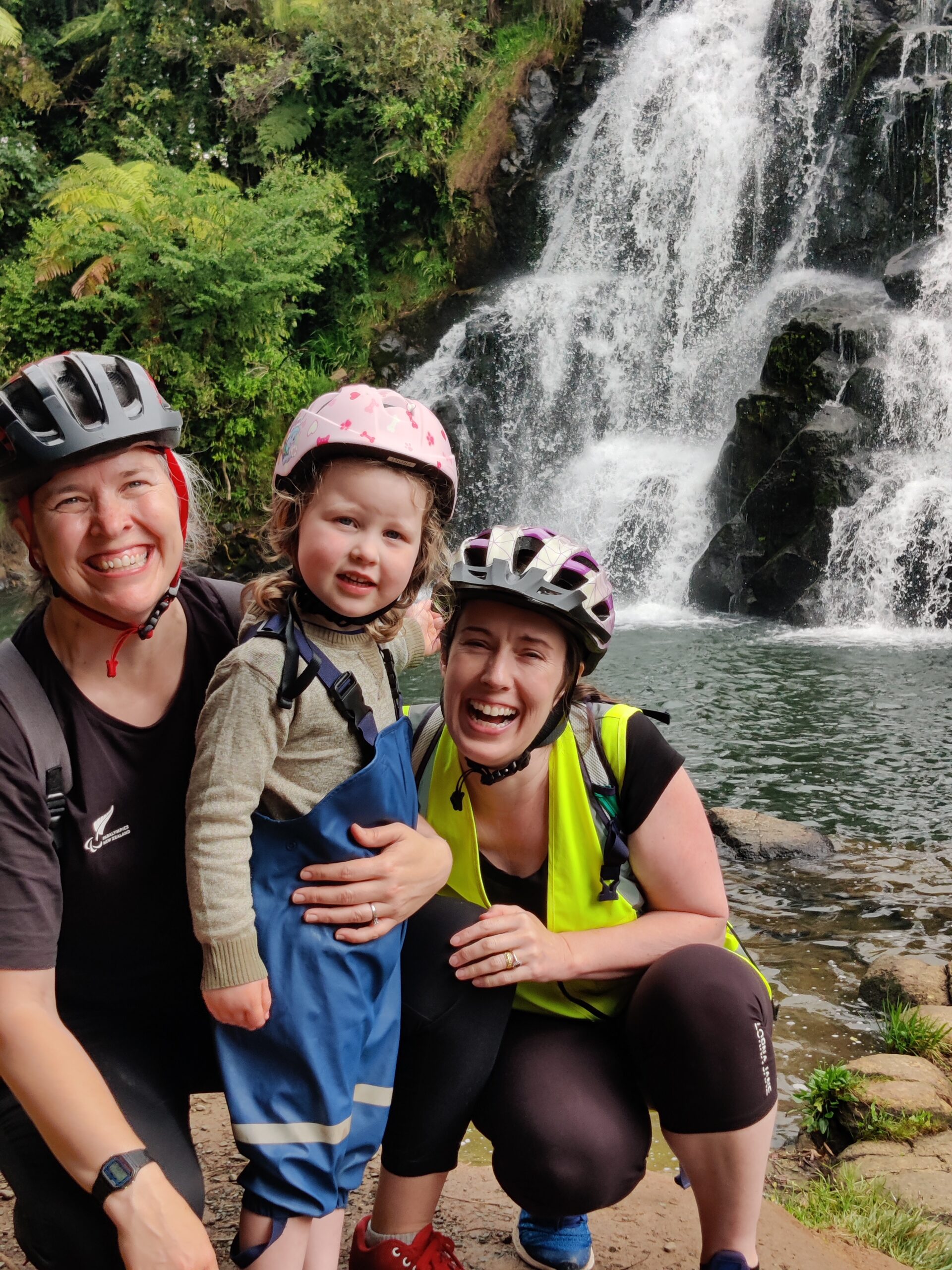 A family with two mums and a toddler smile in front of a waterfall. They are all wearing bike helmets.