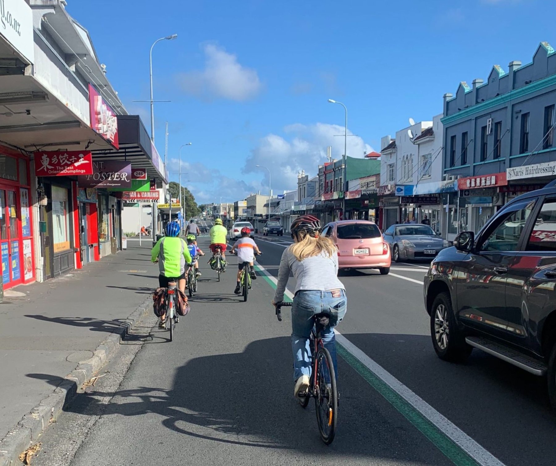 adults and kids cycle on a painted cycleway with cars alongside on a busy city street on a sunny day