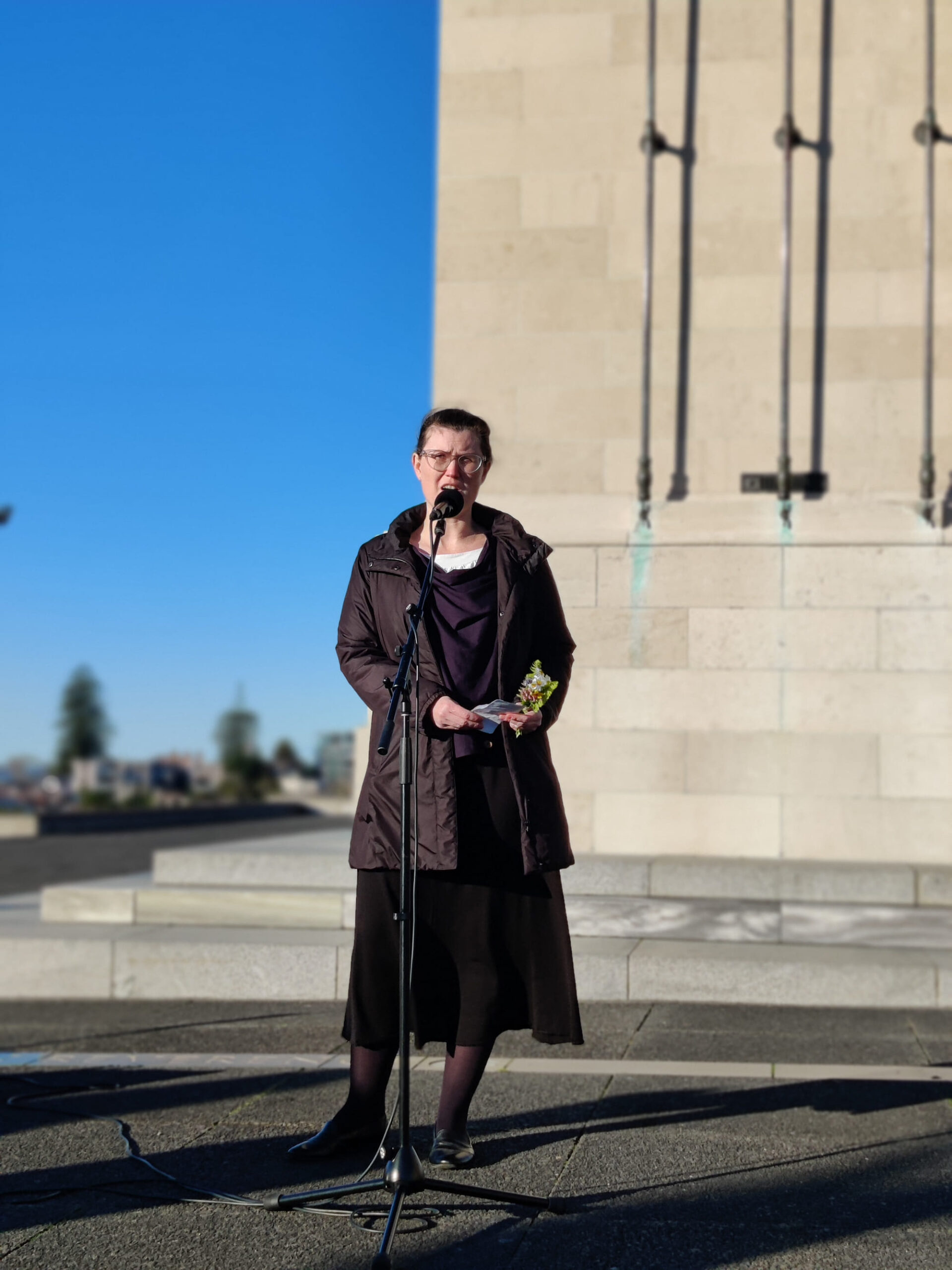A woman in black speaks into a microphone in front of a cenotaph