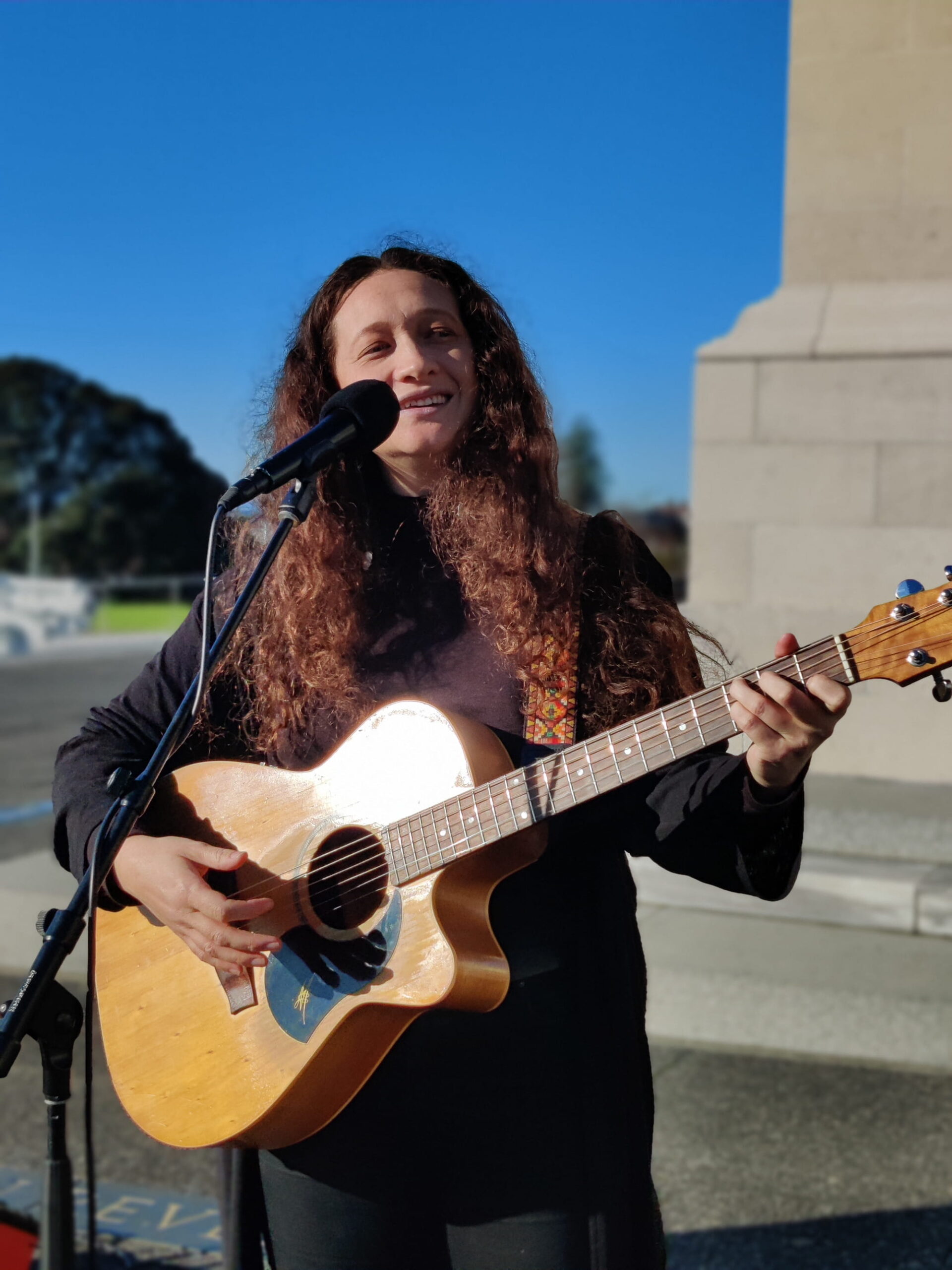 A singer with long curly hair plays a guitar and sings into a microphone in front of a cenotaph