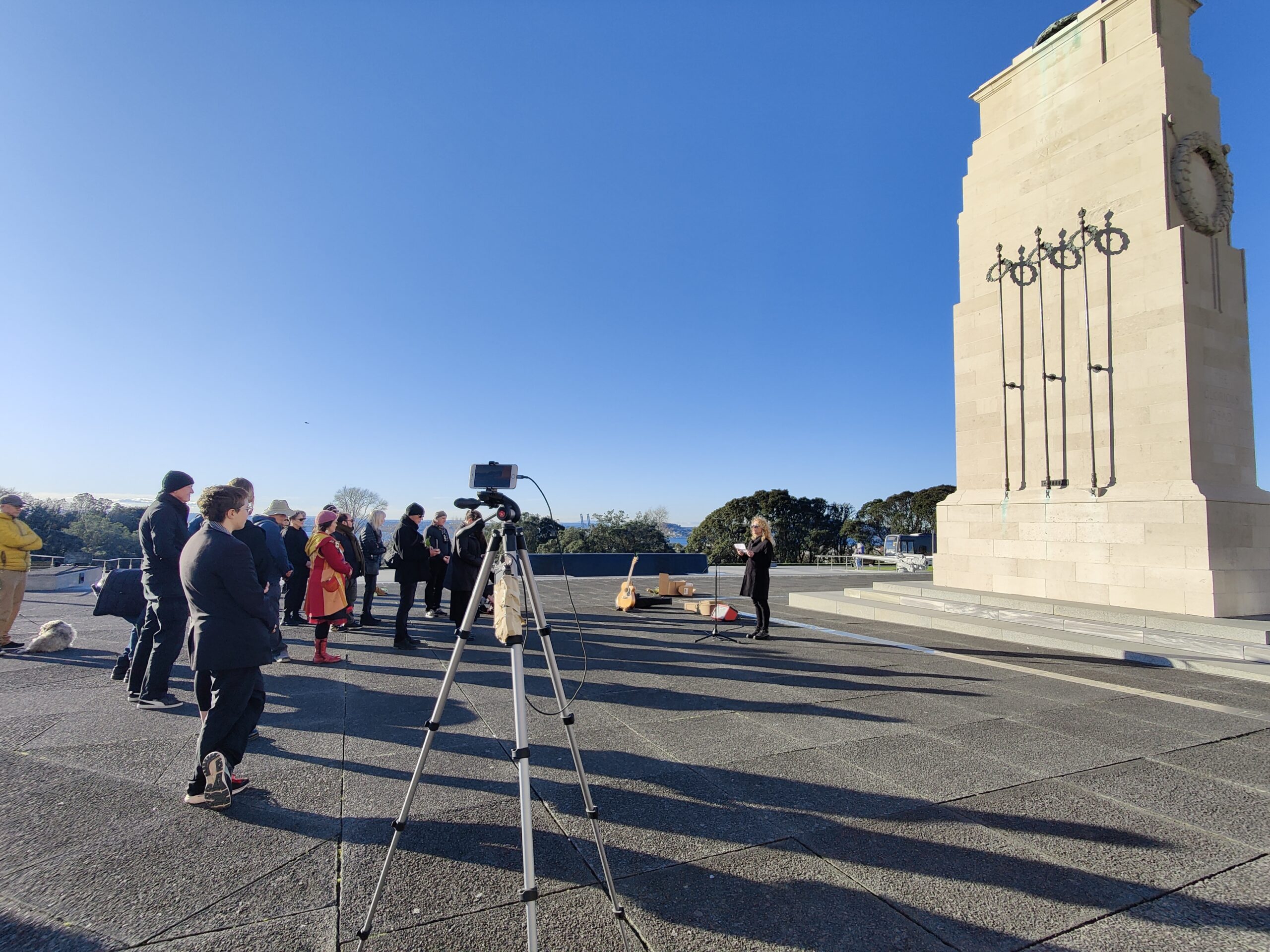 A group of around 25 people listen to a woman speaking into a microphone in front of a cenotaph