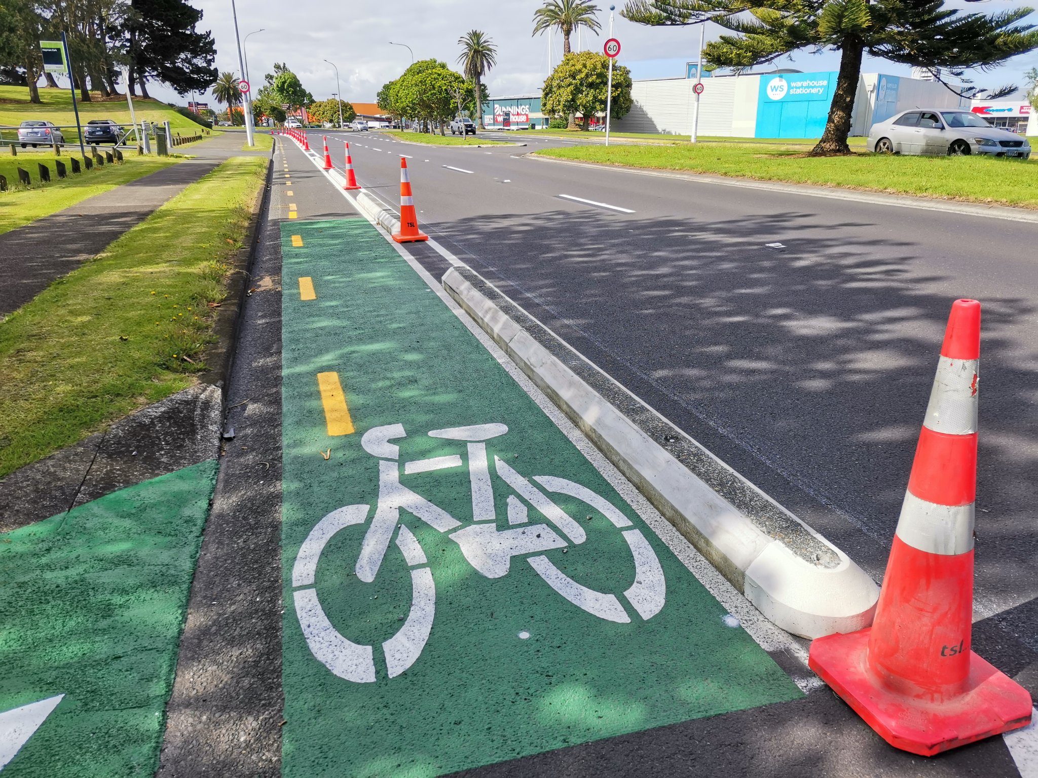 Photo: an on-road unidirectional cycle lane. It is protected from the traffic lane by concrete "biscuit"-style separators, supplemented temporarily by orange road cones. 