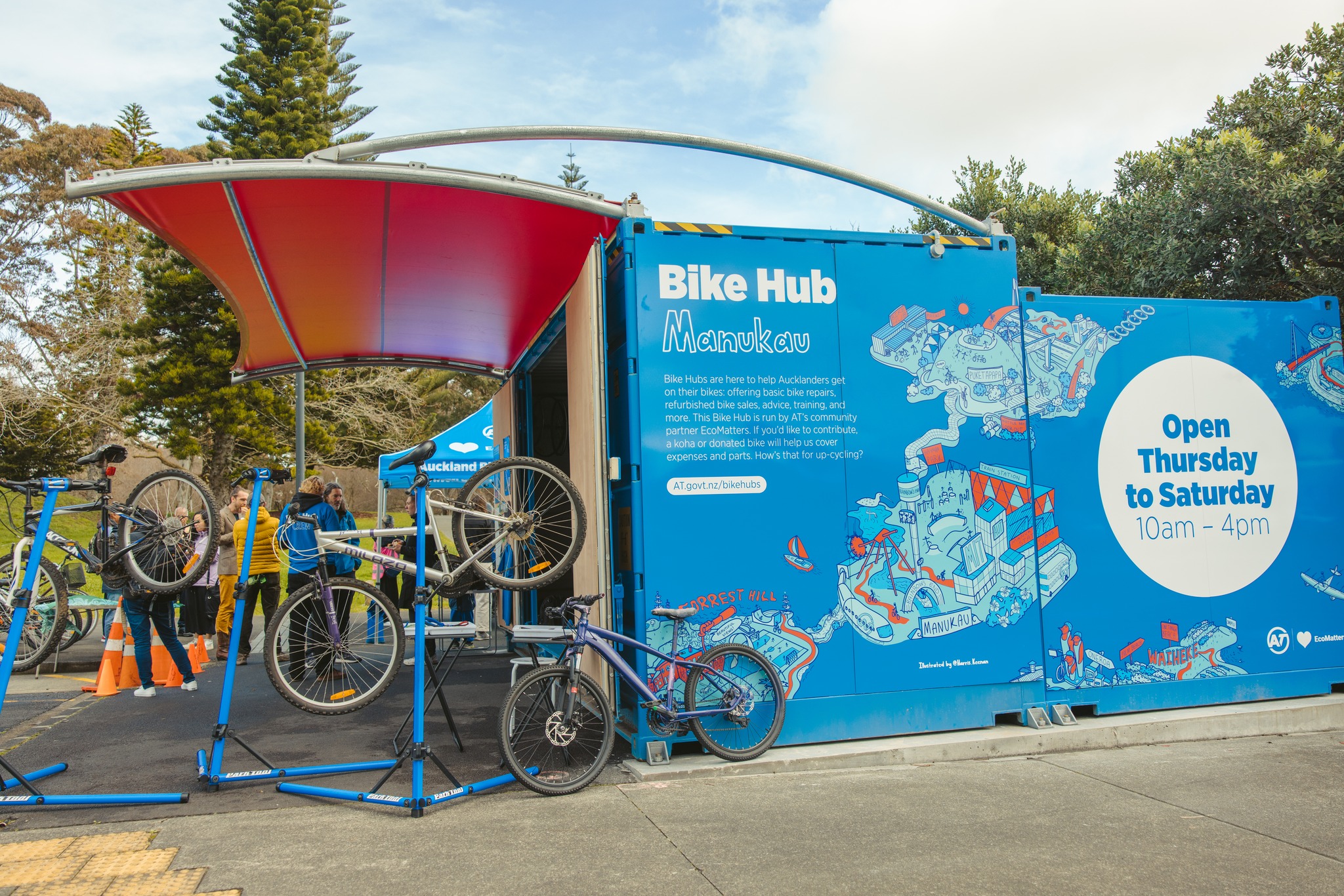 Photo: Manukau Bike Hub's decorated container, as well as a number of bikes on repair stands. In the background, people are standing around talking. 