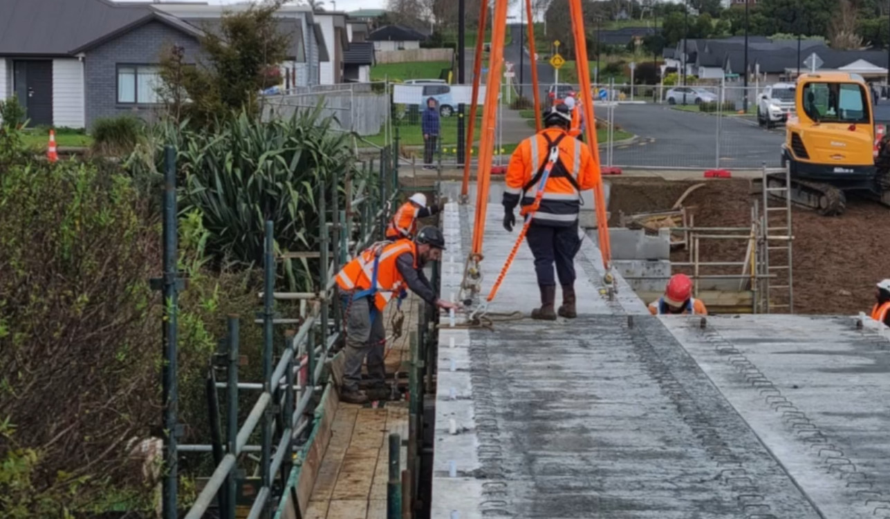 Photo: Construction of a bridge, featuring heavy equipment and scaffolding. Multiple workers in high-vis are pictured. 