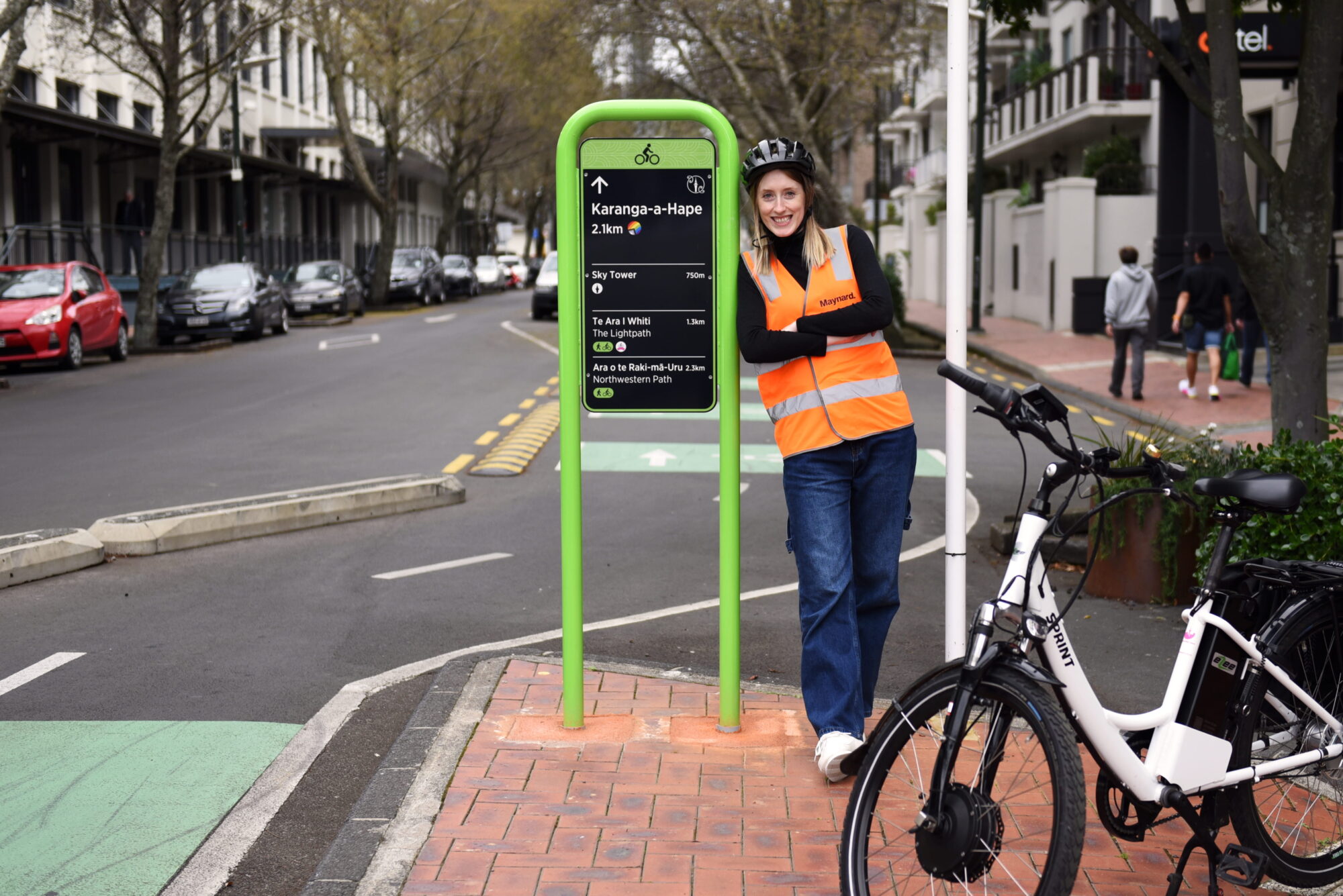 Emme leans against a cycleway sign in the city centre, with her bike parked in front of her