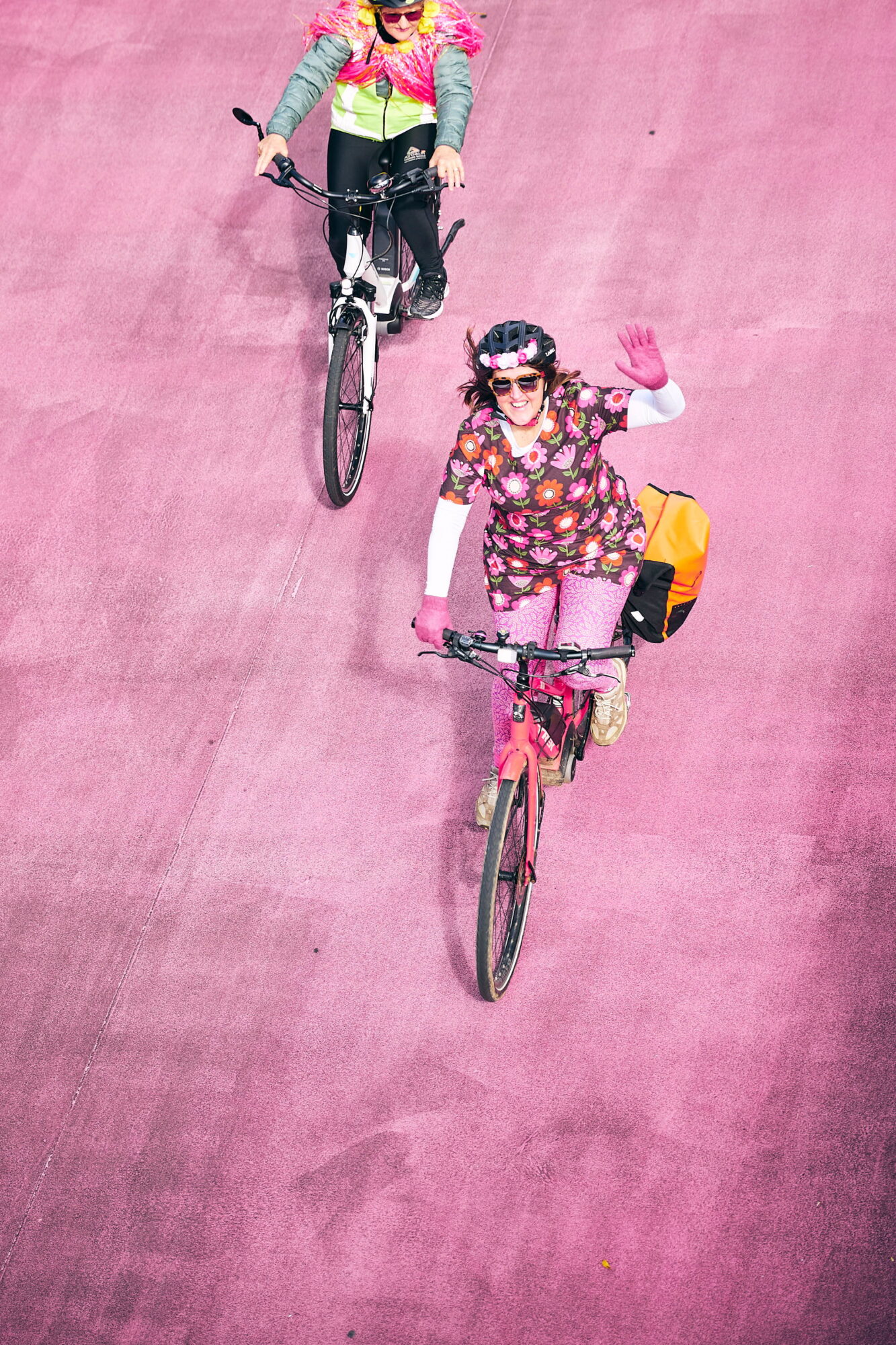 Pink path seen from above, two colourful people on bikes wave up to the camera