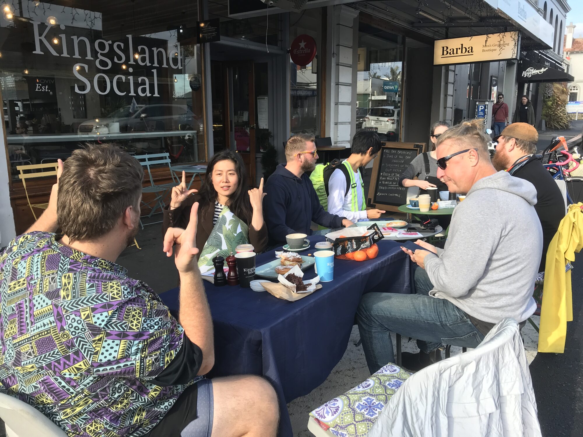 A dining table is set up in a parking space on an urban street. Angie is one of several people enjoying a picnic.