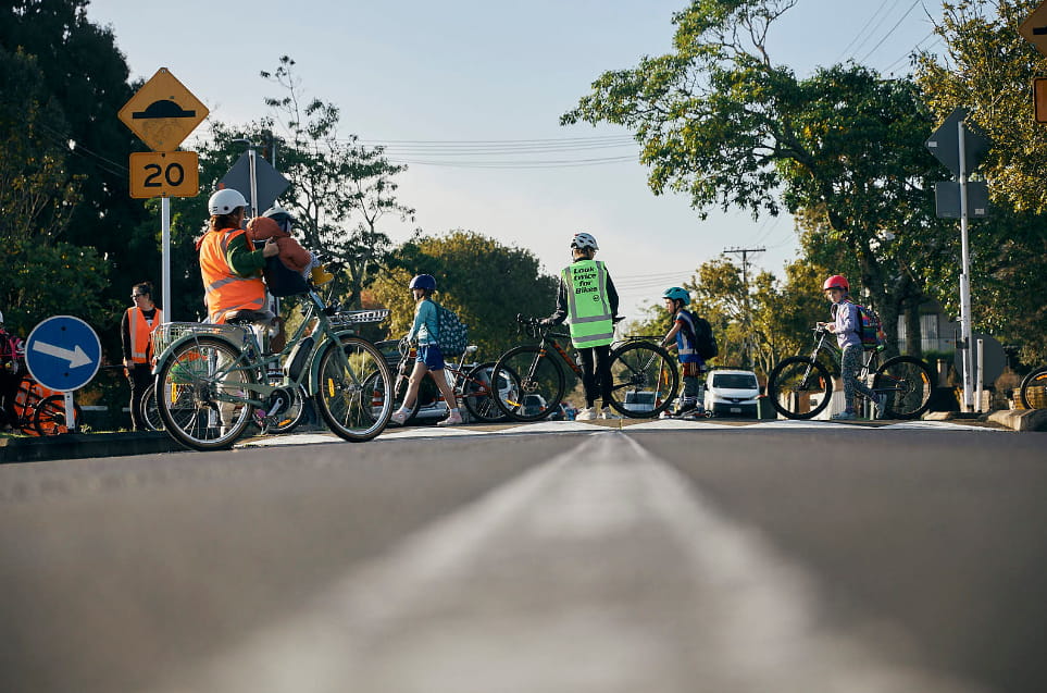 Adult guides stand in the road to stop traffic while kids with bikes cross a zebra crossing.