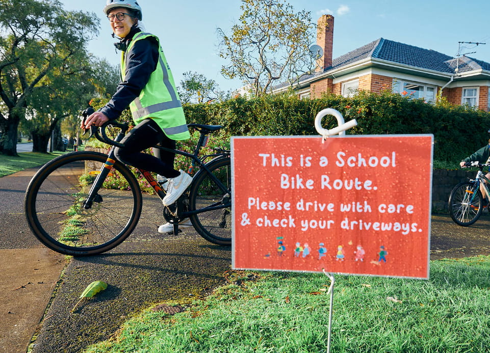 An adult guide on a bike by a sign saying "This is a School Bike Route. Please drive with care and check your driveways."