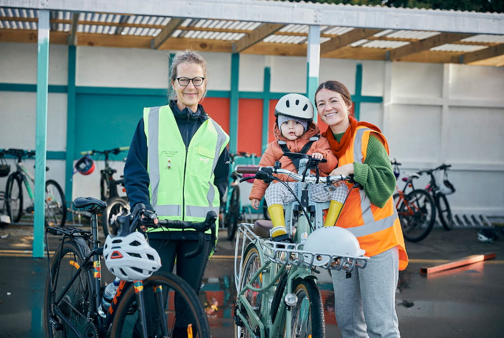 Two women in high vis bike gear stand in front of a bike shed, one woman has a baby on her bike.