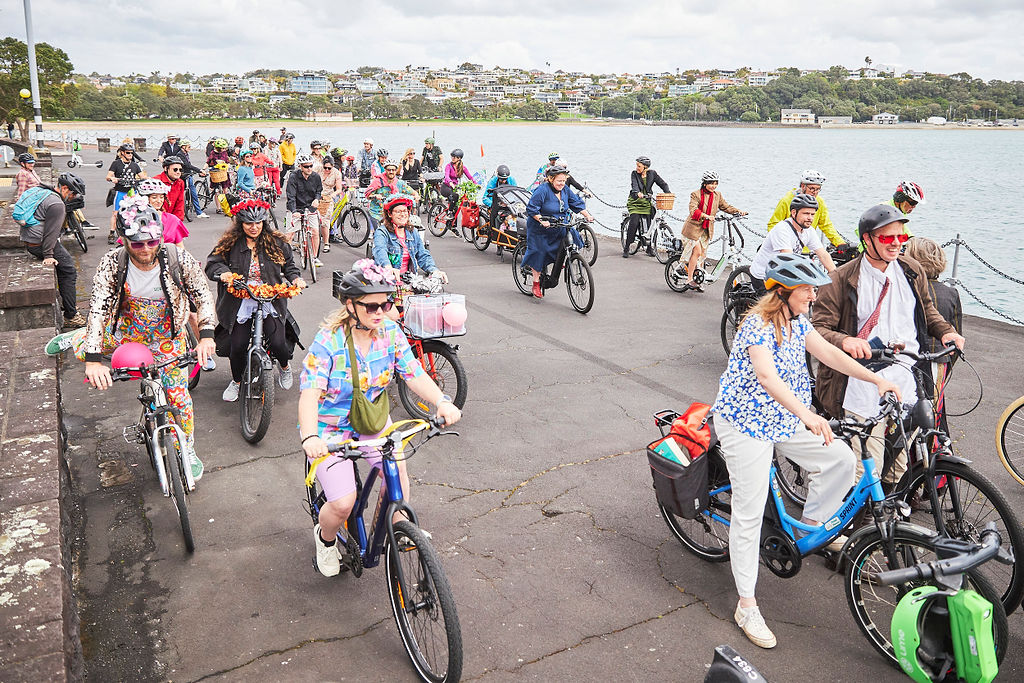 A large colourful group of people on bikes on a path beside the sea.