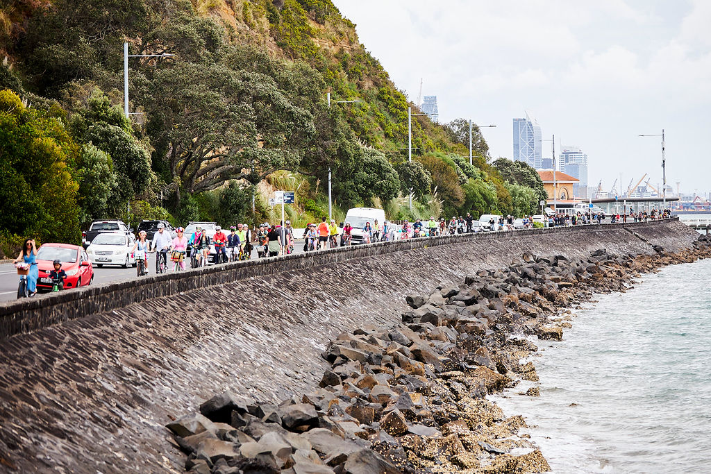 A long colourful line of people on bikes ride along a coastal road