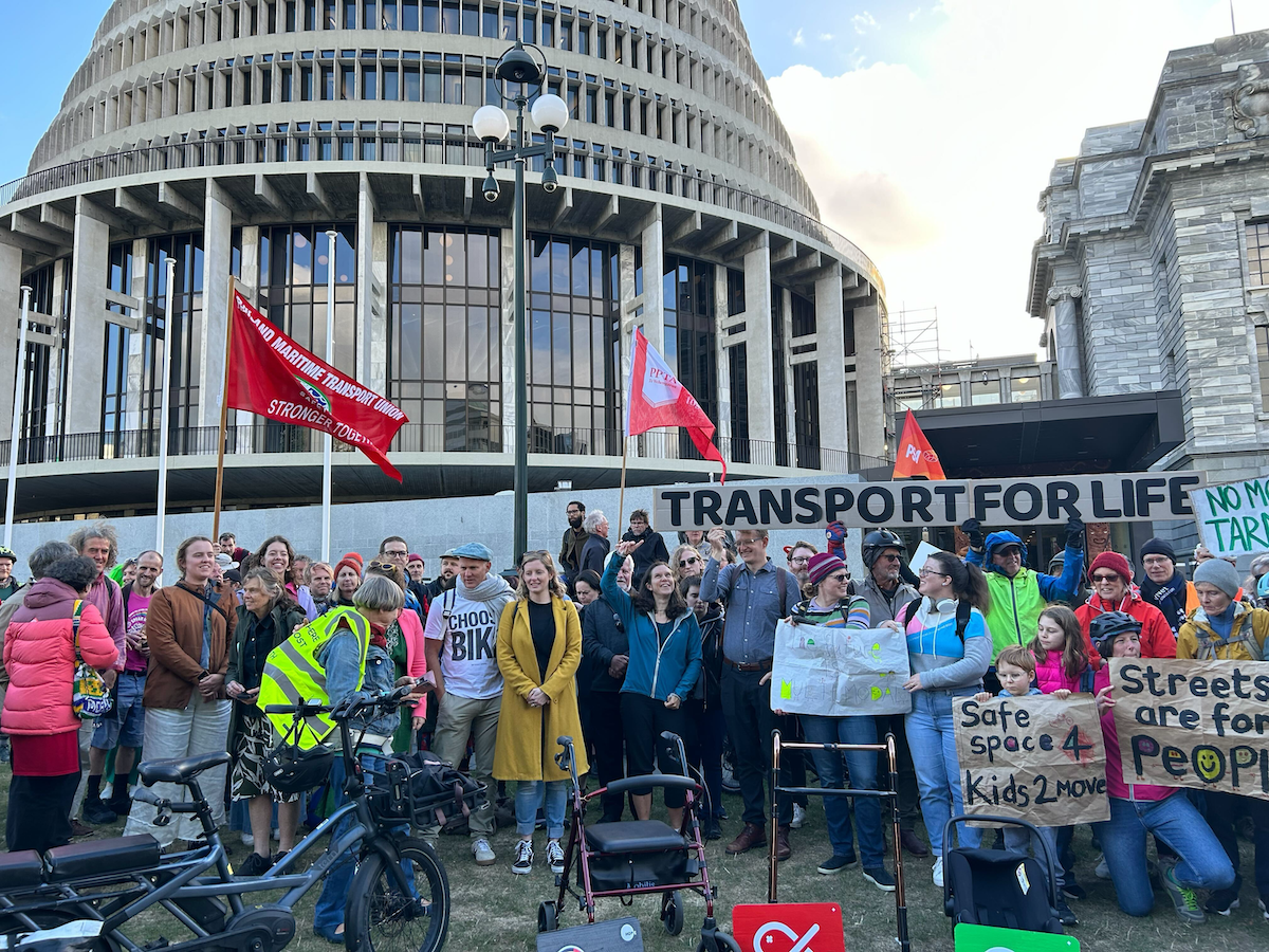 A crowd is holding an array of pro-active transport signs, with bikes and mobility aids in the foreground. The Beehive (NZ parliament building) is in the background.