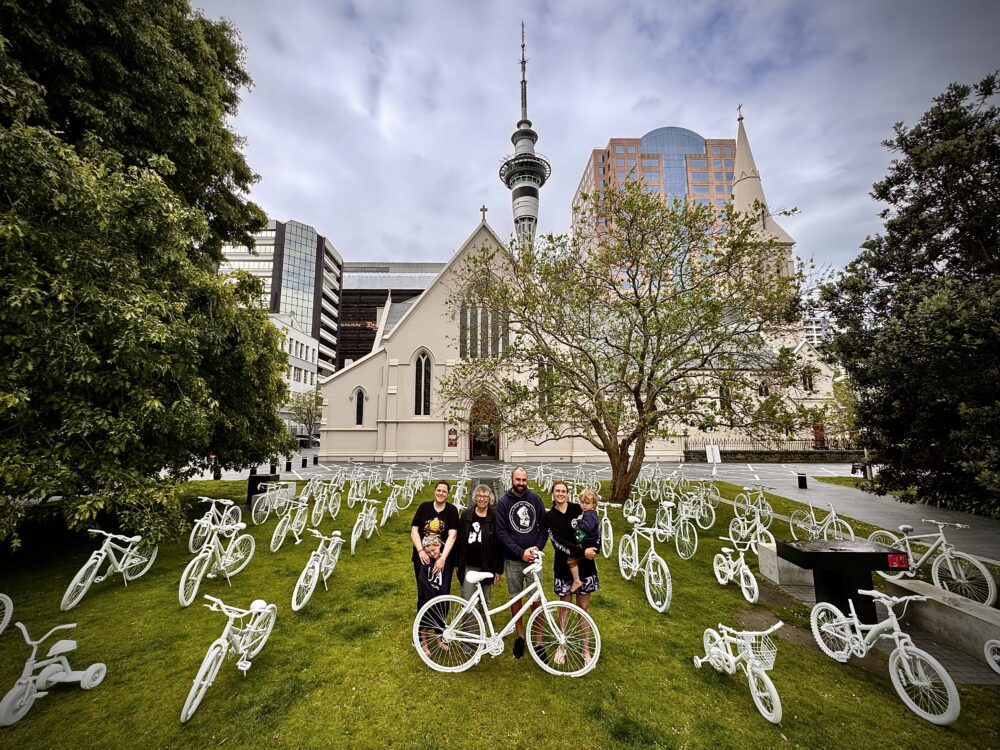 A family stands amid an array of many ghost bikes on grass in front of a church